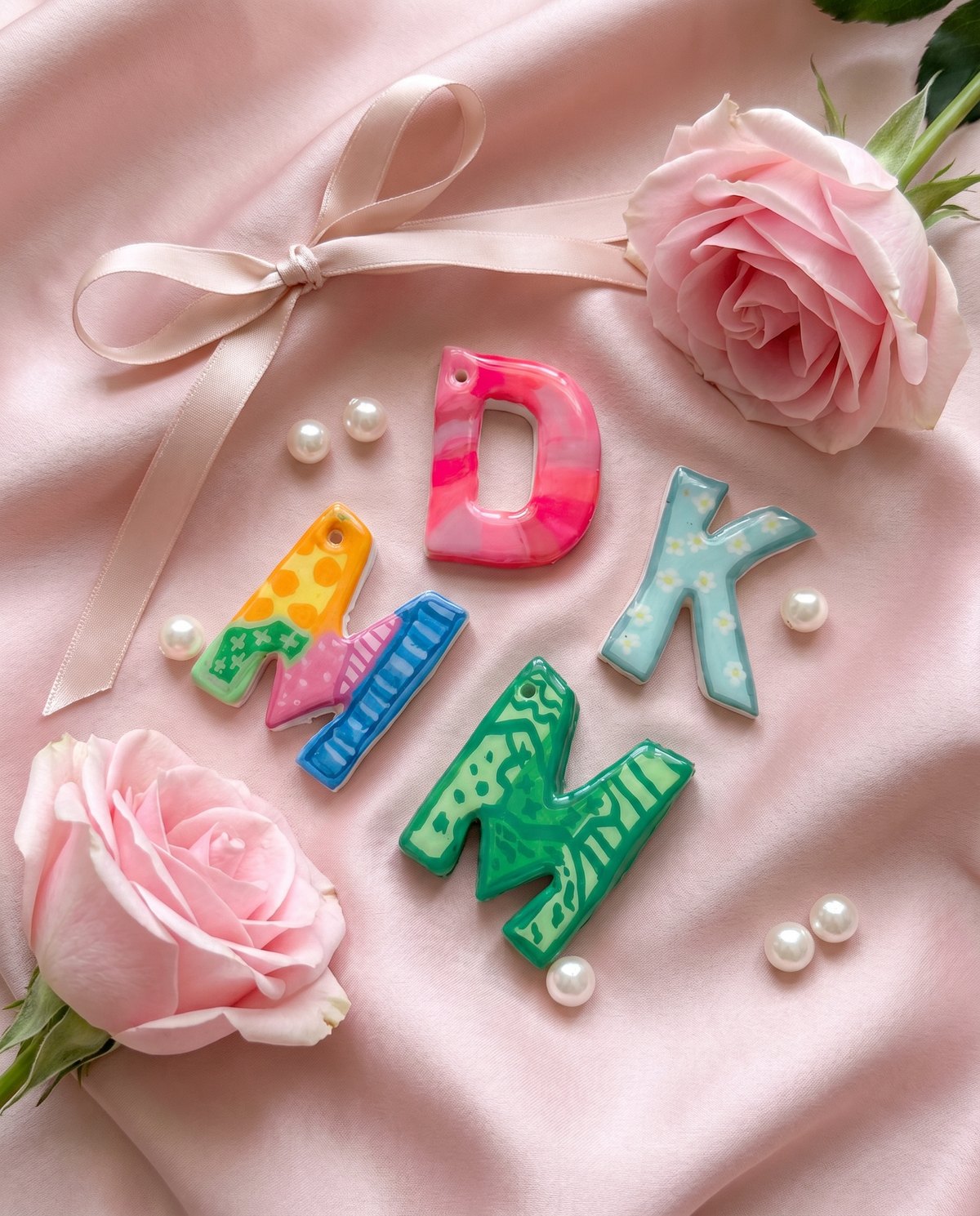 Four hand-painted letter charms styled on blush silk with roses and pearls.