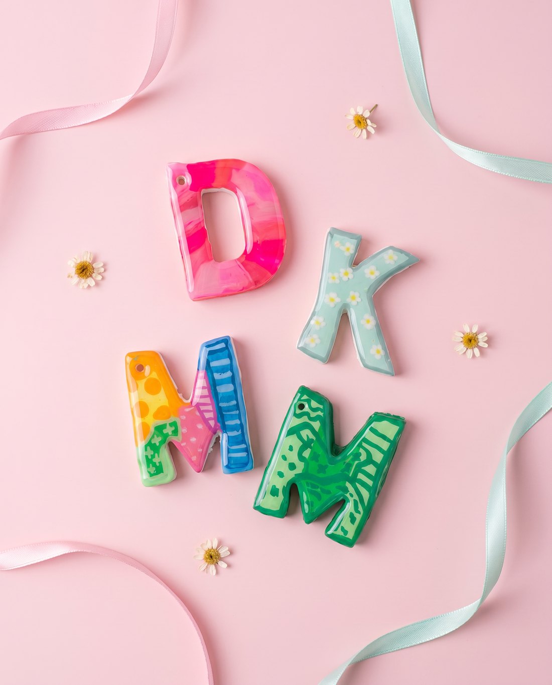 Flat-lay of four colourful hand-painted letter charms on a pink background.