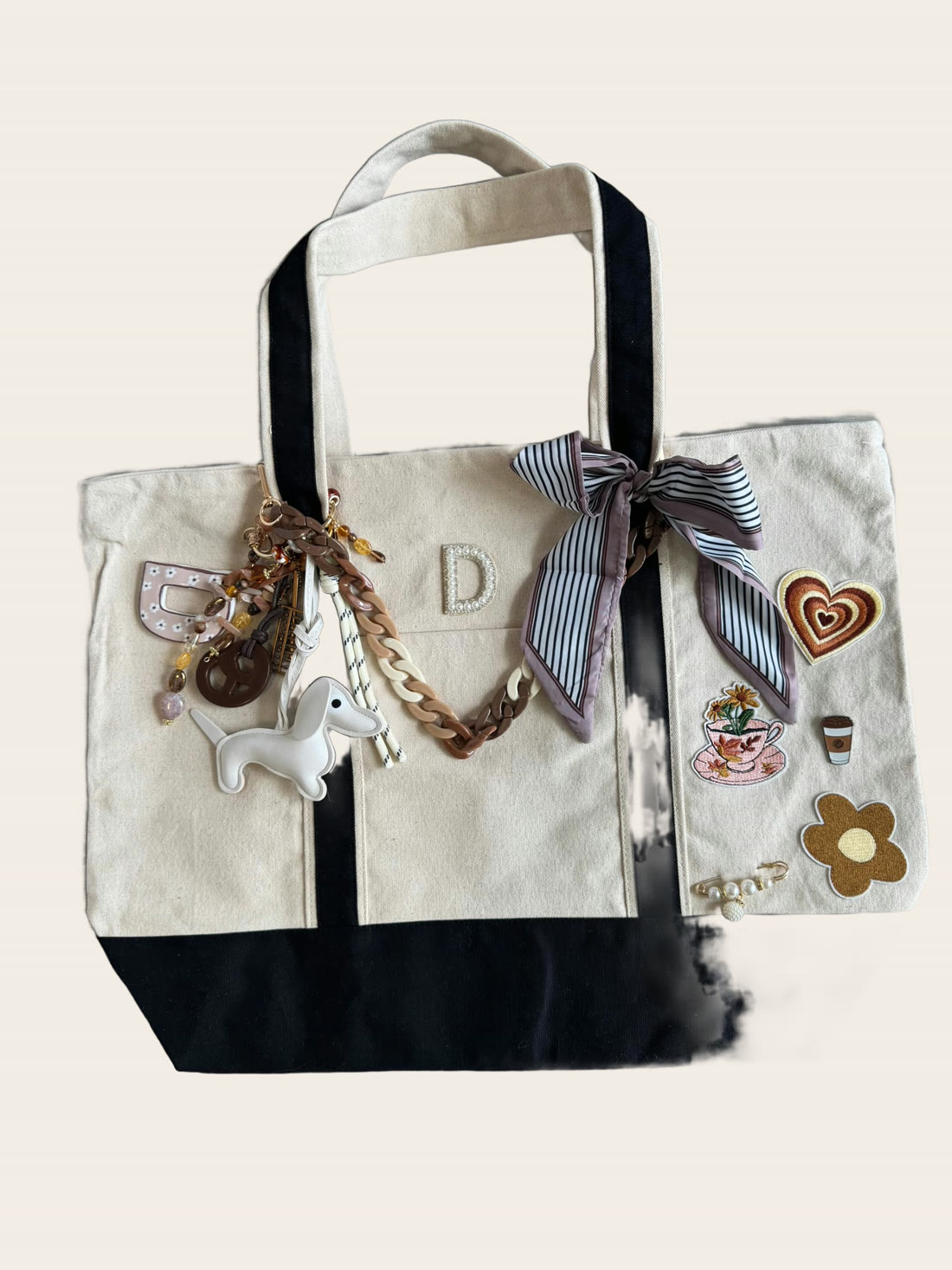 A cream and black canvas tote with a pearl initial D, a dog charm, heart and flower patches and a striped twilly.