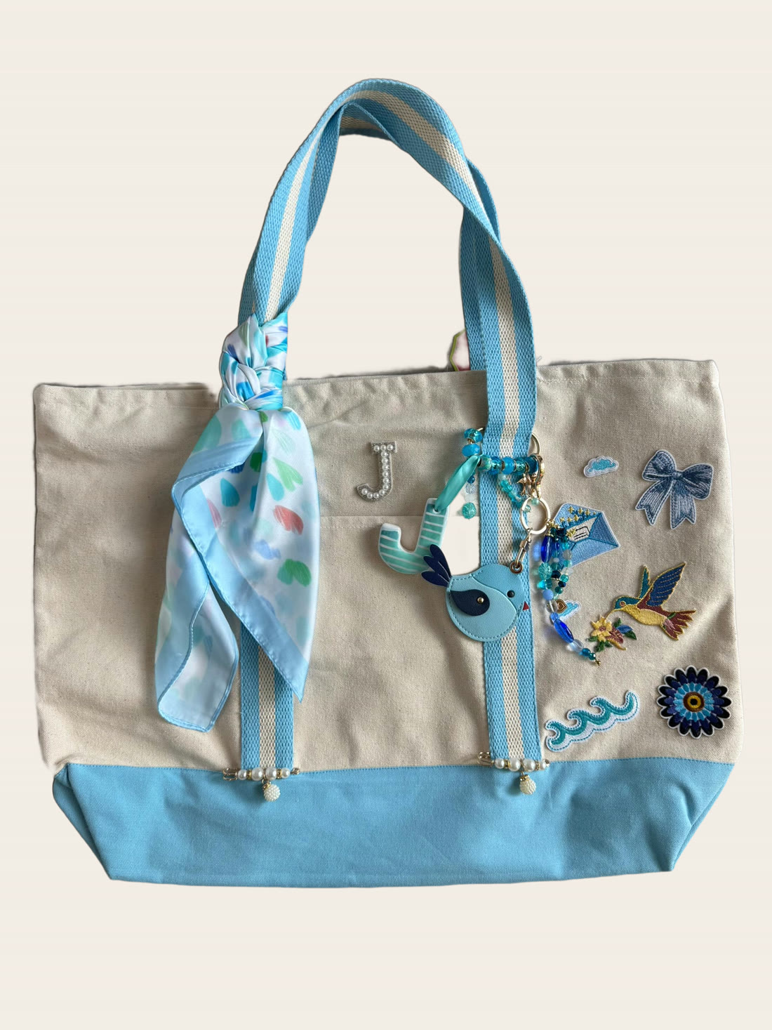 A powder-blue canvas tote with an initial J patch, a bluebird charm and pearls on the handle.