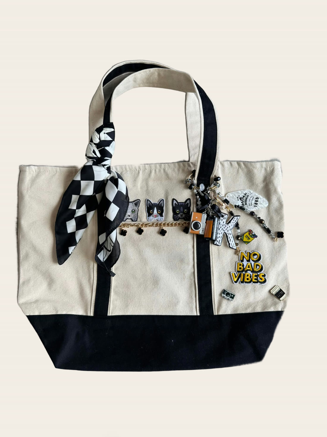 A cream and black canvas tote with cat patches, a checkered twilly, beaded charms and NO BAD VIBES lettering.