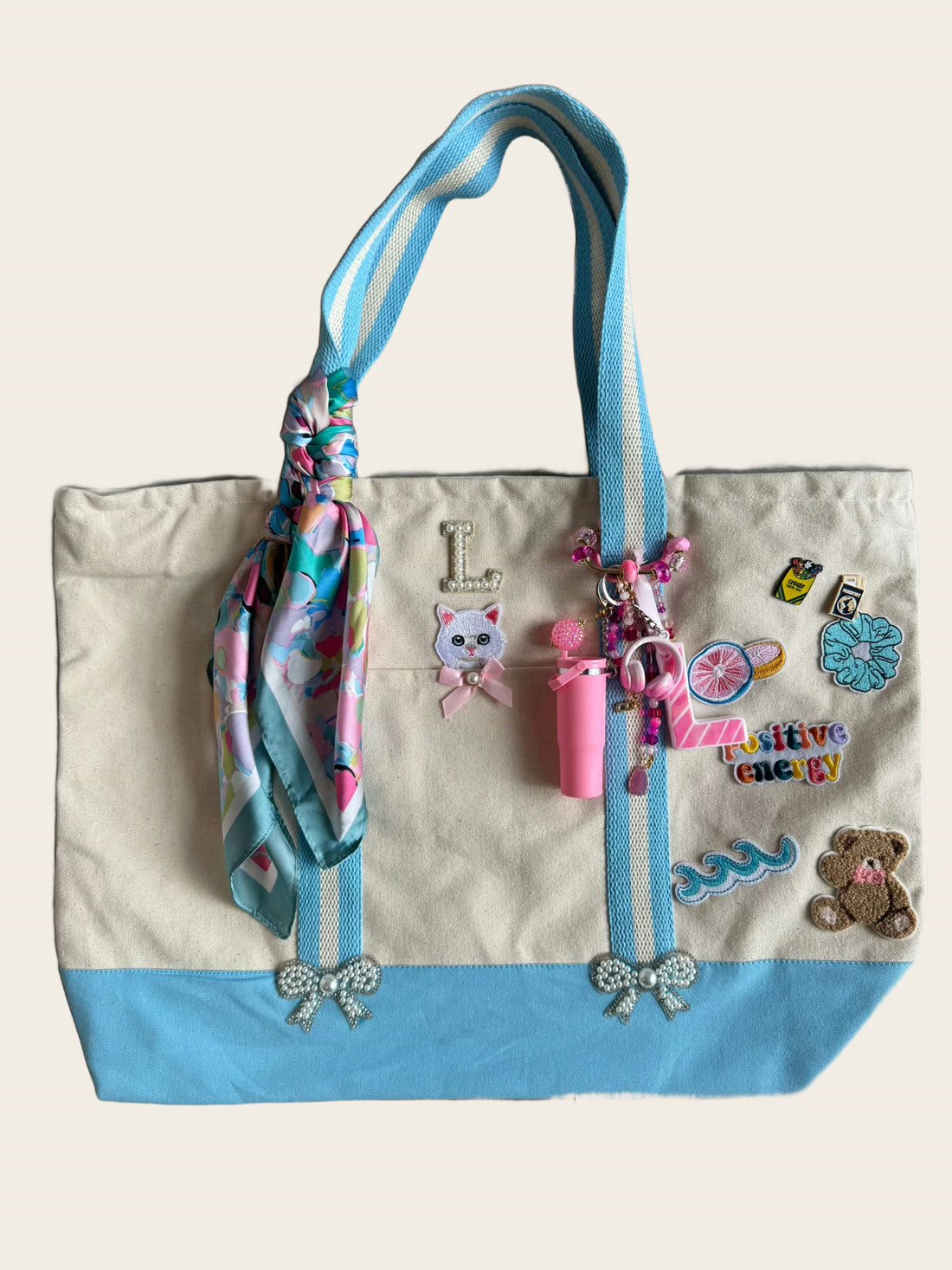 A cream and powder-blue canvas tote with a cat initial L, a teddy bear, positive energy patch and a floral silk scarf.